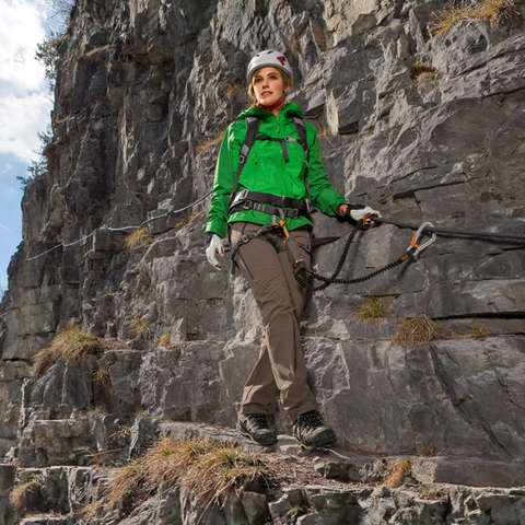Eine Frau geht in Bergstiefeln und grüner Softshelljacke einen schmalen Gebirgspass entlang.