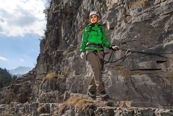 Eine Frau geht in Bergstiefeln und gr&uuml;ner Softshelljacke einen schmalen Gebirgspass entlang.