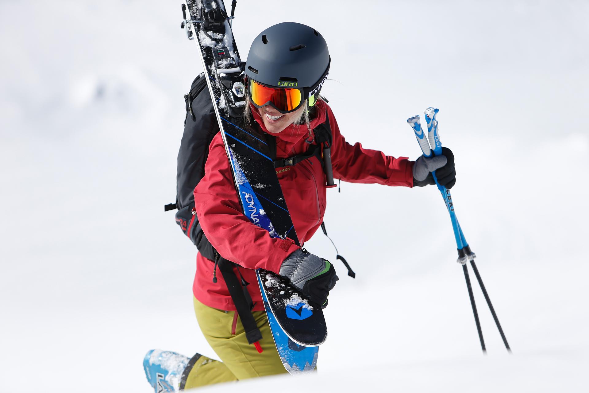 Eine Skifahrerin geht mit ihren Ski auf der Schulter und Skist&ouml;cken in der Hand ein St&uuml;ck die Piste hinauf