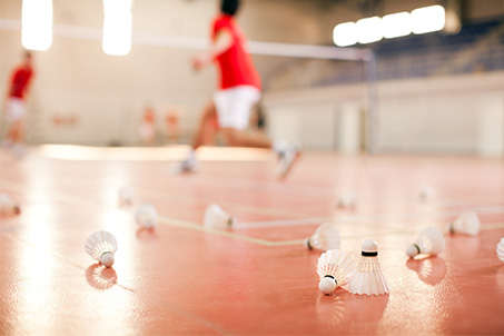 Diverse Badmintonbälle liegen auf dem roten Hallenboden. Im Hintergrund wird ein Badminton Match ausgetragen.
