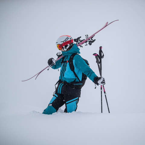 Eine Skifahrerin geht mit ihren Ski auf der Schulter und Skistöcken in der Hand durch kniehohen Tiefschnee.