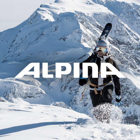 Ein Mann und eine Frau stehen vor einem Bergpanorama. Sie tragen beide verspiegelte Alpina Skibrillen und Skihelme.