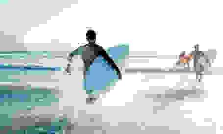 Mehrere Surfer laufen in Neoprenanzug und Surfbrett unter dem Arm ins Meer.