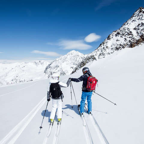 Ein Skifahrer und eine Skifahrerin fahren durch flaches Gelände im Tiefschnee.