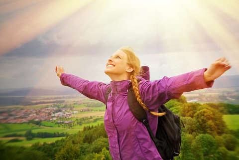 Eine Frau steht auf einem Berg und trägt eine pinke Regenjacke. Im Hintergrund ist die Sonne zu sehen und die Frau streckt die Arme in den Himmel.