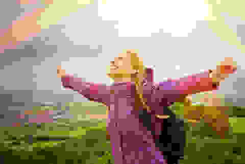 Eine Frau steht auf einem Berg und trägt eine pinke Regenjacke. Im Hintergrund ist die Sonne zu sehen und die Frau streckt die Arme in den Himmel.