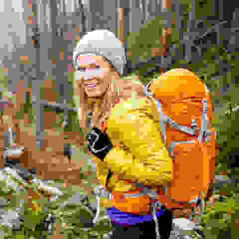 Eine Frau wandert in orange farbener Thermojacke durch den Wald. Auf dem Rücken trägt sie einen großen Wanderrucksack.