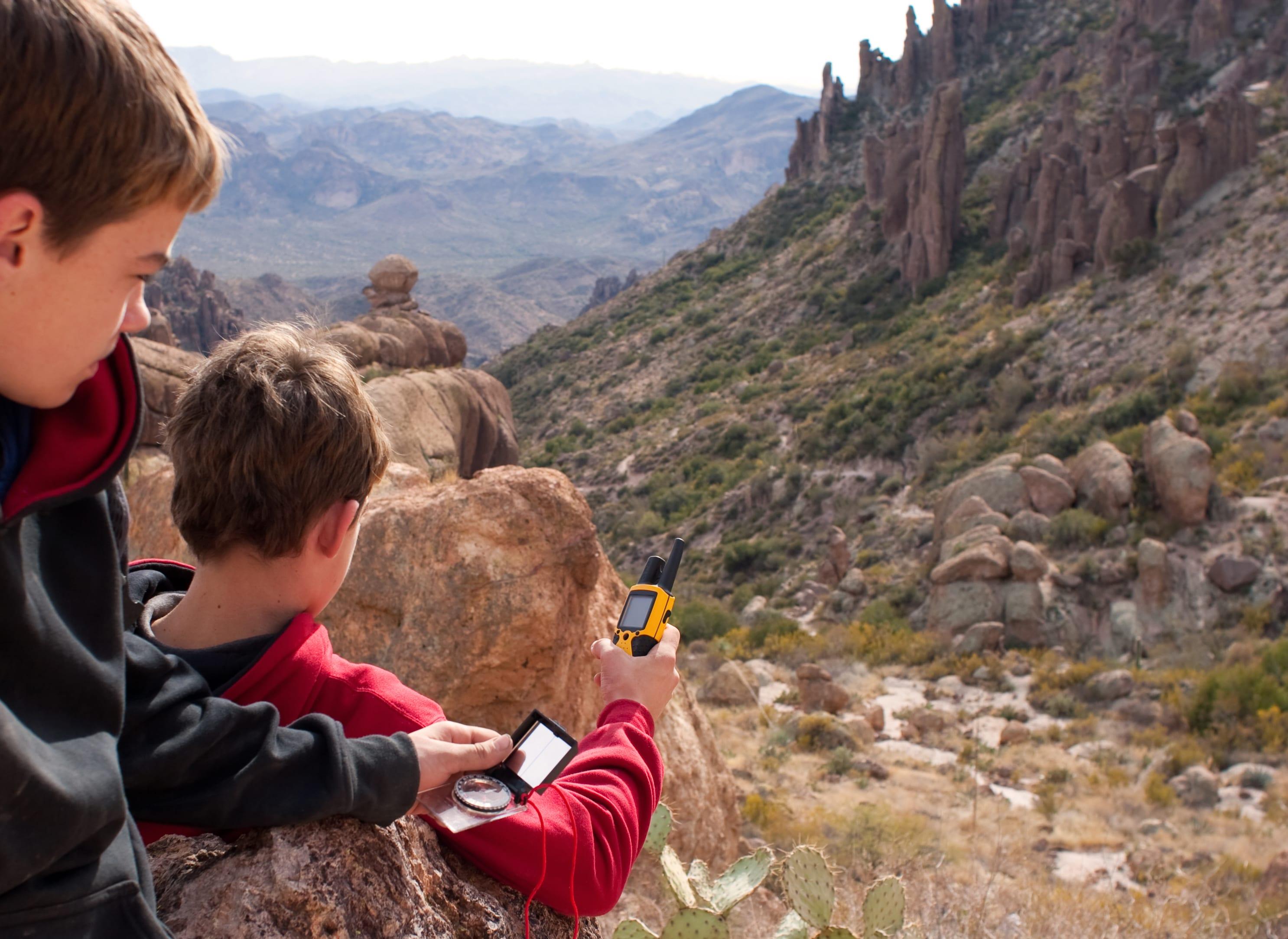 Zwei Jungen befinden sich in einer steinigen Natur und halten ein GPS-Ger&auml;t in der Hand