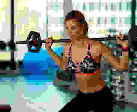 Eine Frau trainiert in schwarzer Trainingshose und Sport-BH mit einer Langhantel in einem Fitnessstudio.