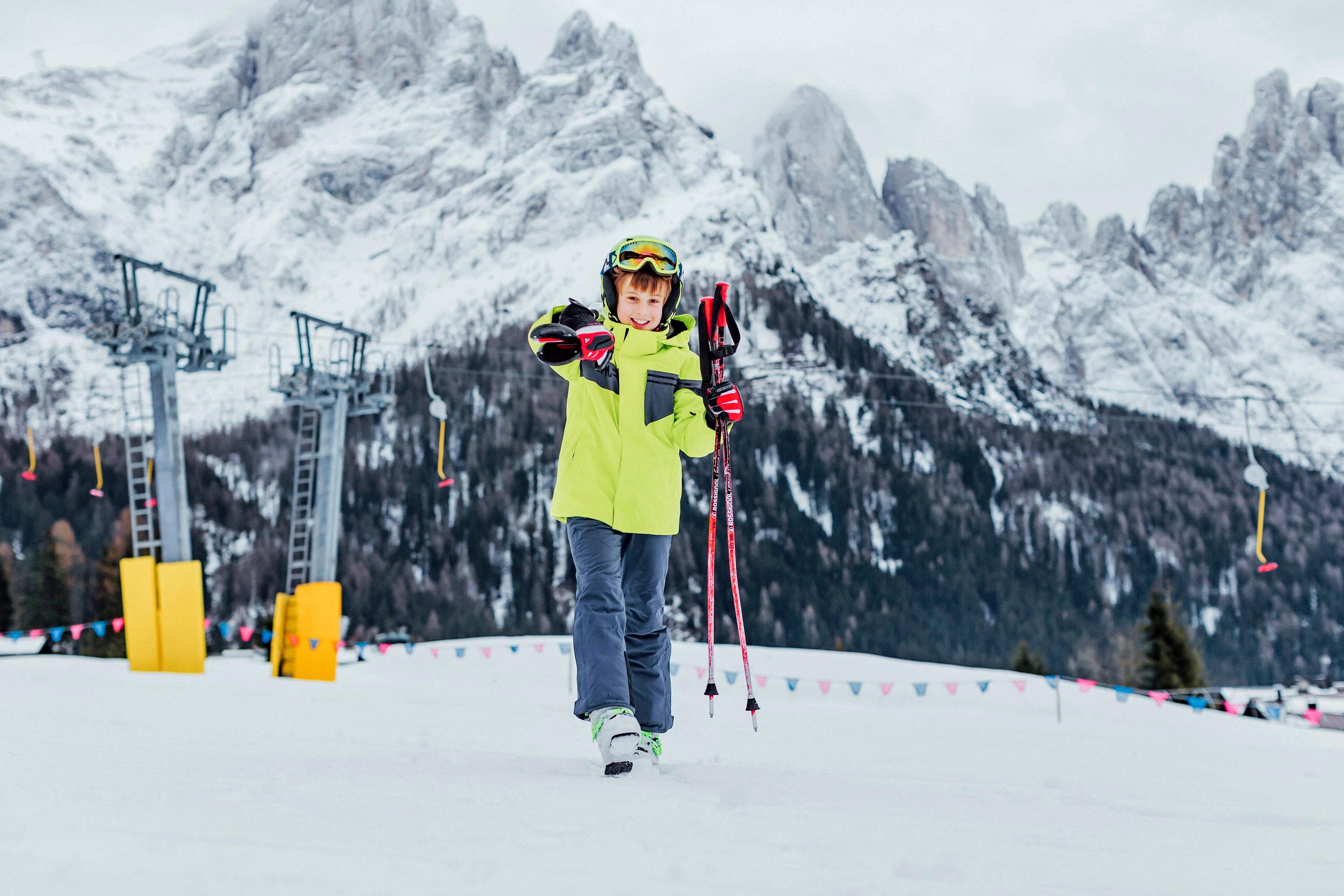 Ein Kind steht mit Ski Ausr&uuml;stung und den Skiern &uuml;ber der Schulter auf einer roten Piste.