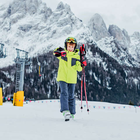 Ein Kind steht mit Ski Ausrüstung und den Skiern über der Schulter auf einer roten Piste.