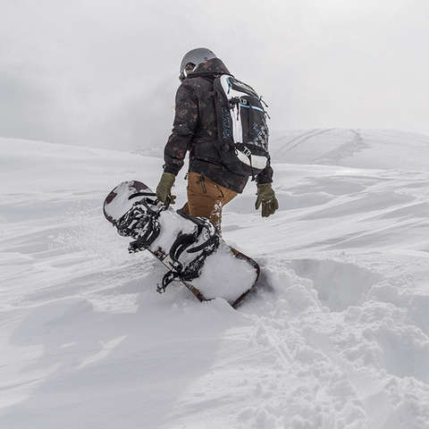 Ein Snowboarder trägt sein Snowboard einen kleinen Tiefschneehang hinauf.