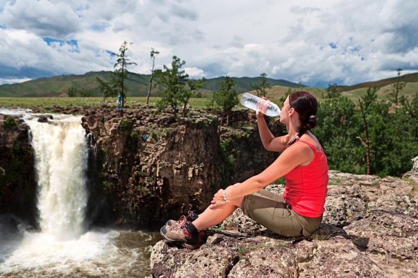 Eine Frau sitzt in der Natur auf einem Felsen und trinkt Wasser aus einer Wasserflasche
