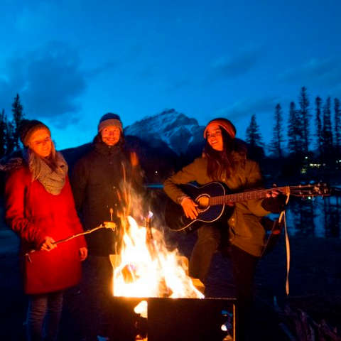 Eine Gruppe von Leuten steht am Abend um ein Lagerfeuer herum. Eine Frau spielt dazu Gitarre.