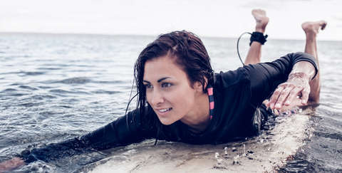 Eine Frau liegt im Wasser auf ihrem Surfbrett und paddelt mit den Armen. Sie trägt einen Surfanzug aus Lycra.