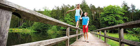 Ein Mann und eine Frau gehen über eine Holzbrücke. Die Frau balanciert dabei auf dem Geländer und der Mann hält sie an der Hand um sie zu stützen. Beide tragen Kleidung von Maier Sports.