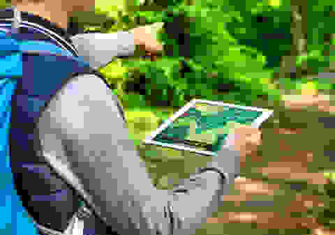 Ein Mann hält ein Tablet in der Hand. Darauf zu sehen ist eine geöffnete Navigationsapp, die den Weg zum nächsten Cache anzeigt.