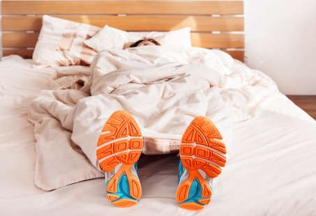Eine Frau liegt im Bett unter einer weißen Decke. Sie trägt Turnschuhe an ihren Füßen, die unter der Decke herausragen.