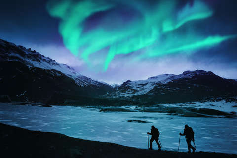 Zwei Nachtwanderer laufen an einem Fjord entlang während am Himmel Polarlichter leuchten
