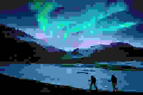 Zwei Nachtwanderer laufen an einem Fjord entlang während am Himmel Polarlichter leuchten