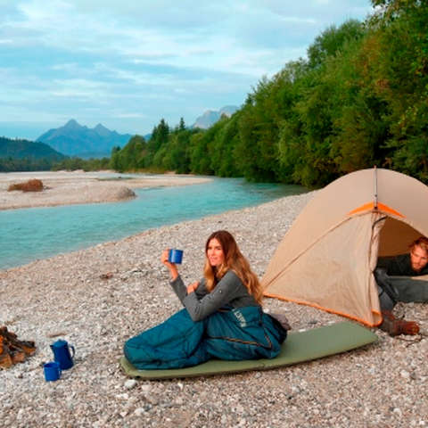 Ein Mann und eine Frau zelten am Ufer eines Sees. Der Mann schaut aus dem Zelt und die Frau sitzt davor auf einem Primaloft Schlafsack. 