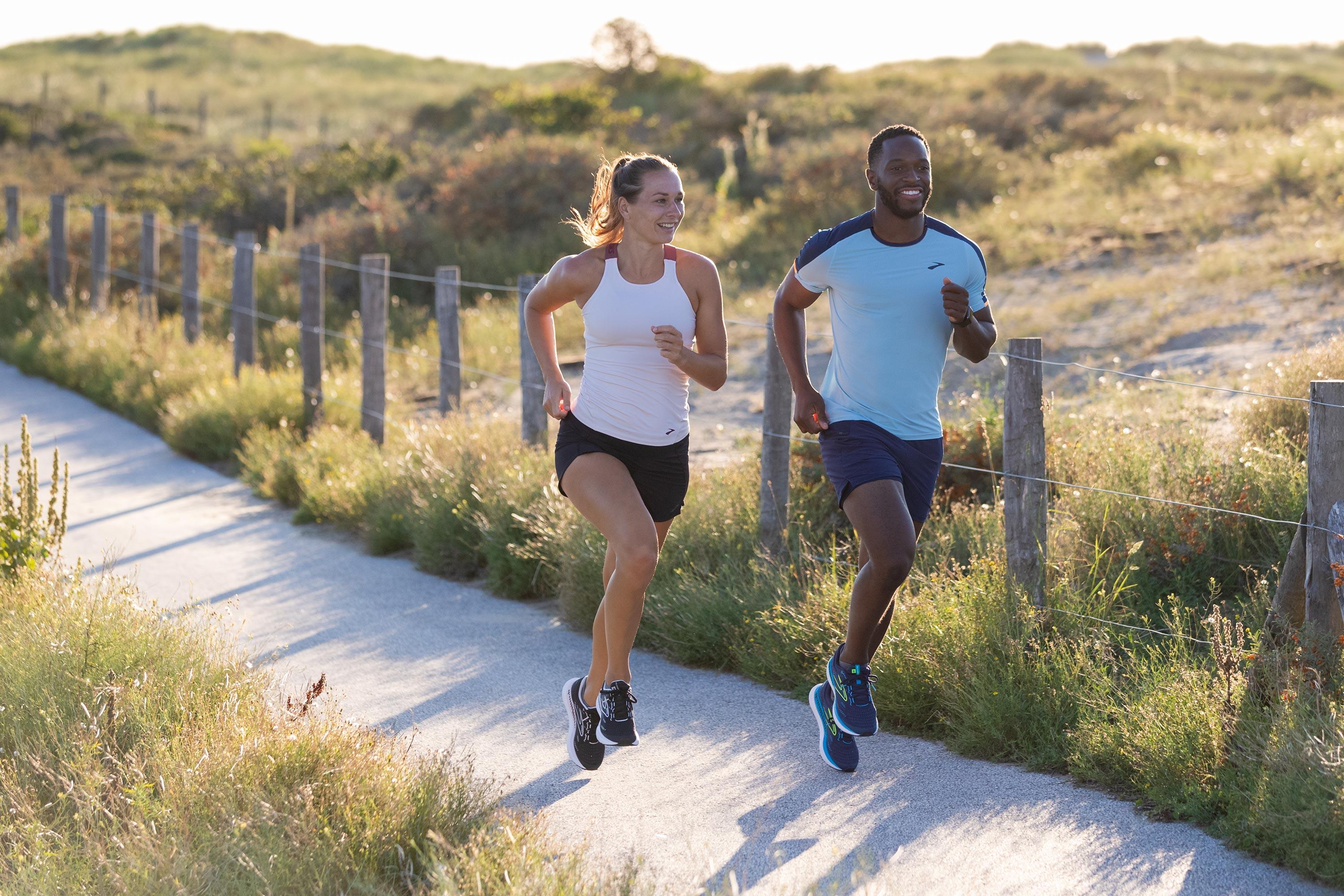 Zwei Jogger laufen nebeneinander durch eine von Tälern eingerahmte Landschaft.