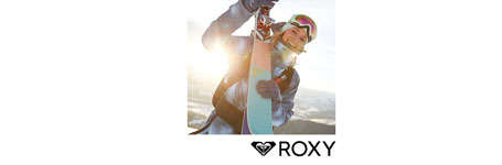 Eine Frau schaut lächelnd in die Kamera. Sie trägt ein komplettes Roxy Skioutfit und hält einen Ski in Richtung Kamera.