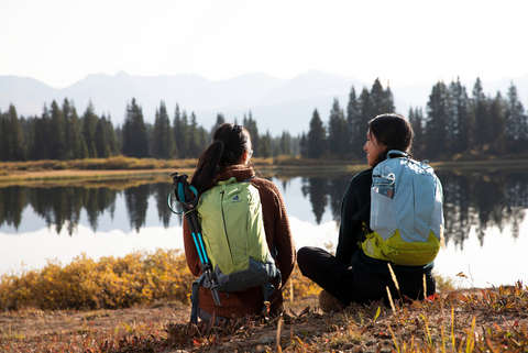 Zwei Personen sitzen am Seeufer mit Rucksäcken auf dem Rücken