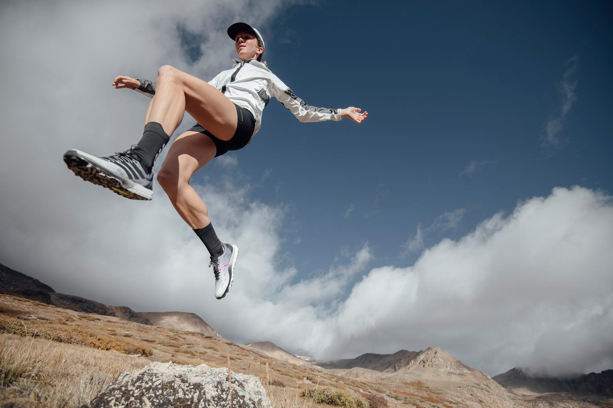Ein L&auml;ufer macht in Adidas-Wanderschuhen einen weiten Sprung &uuml;ber steiniges Gel&auml;nde vor offener Berglandschaft.