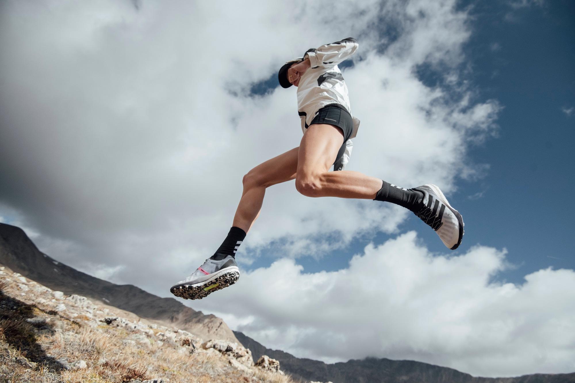 Ein L&auml;ufer springt in Adidas-Schuhen dynamisch &uuml;ber einen Felsen vor Bergkulisse.