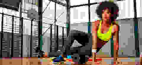 Eine Frau trainiert in einem Fitnessstudio mit einer Faszienrolle. Sie trägt dabei eine blaue Trainingshose und einen neongelben Sport-BH von Nike.