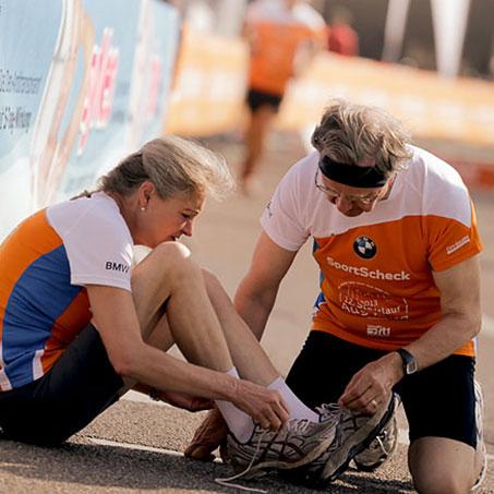Eine Frau sitzt auf dem Boden und bindet ihre Laufschuhe neu. Ein Mann kniet vor ihr um sie dabei zu unterstützen.