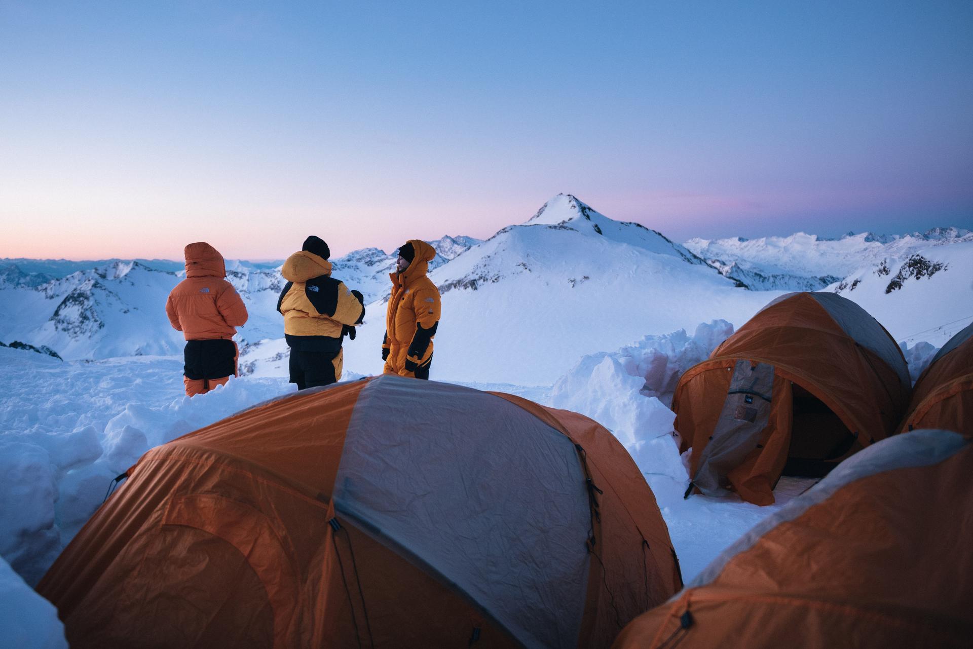 Drei Hochtouren-Geher stehen bei Sonnenuntergang vor dem Eingang der Zelte ihres Hochlagers 