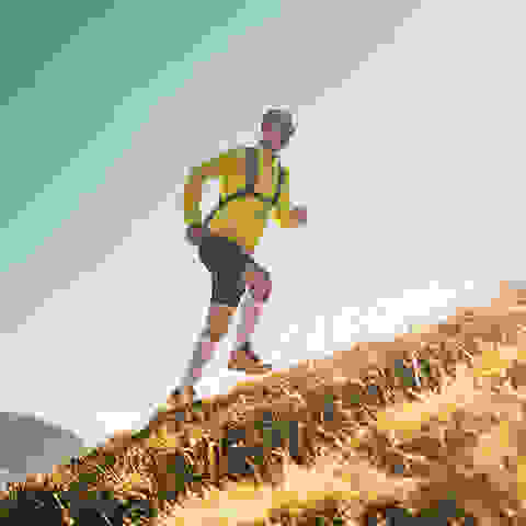 Ein Mann in schwarzer kurzer Hose und gelbem Trainingsshirt läuft beim Trailrunning eine Bergwiese hinauf.