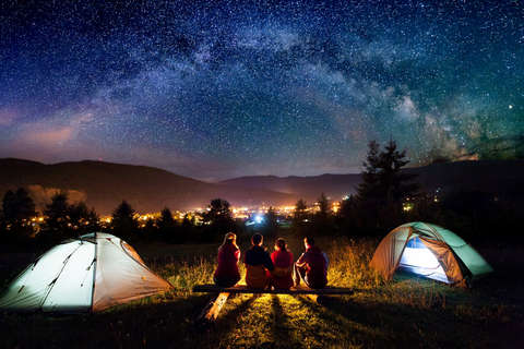 2 Jungs und 2 Mädchen sitzen vor ihrem grünen Zelt und genießen den Sternenhimmel mit Blick auf die Lichter einer Stadt.