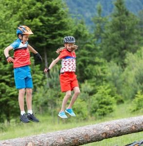 Zwei Kinder mit Fahrradhelmen springen auf einem Ast im Wald