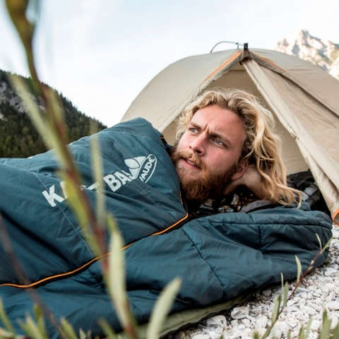 Ein Mann liegt im Schlafsack vor einem geöffneten Zelt.