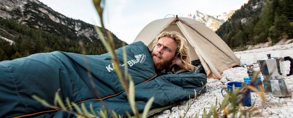 Ein Mann liegt im Schlafsack vor einem ge&ouml;ffneten Zelt.