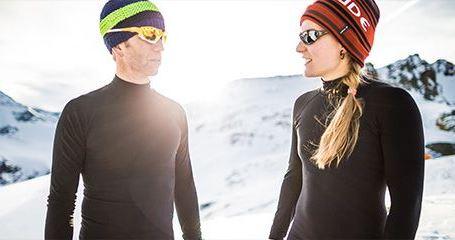 Zwei Personen stehen in Funktionsunterwäsche und Mützen in einer sonnigen Winterlandschaft