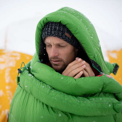 Ein Mann steht in verschneiter Landschaft in einem sehr warmen Schlafsack vor seinem Zelt