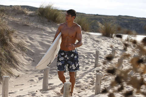 Ein Mann geht mit Badehose und Surfbrett Richtung Strand.