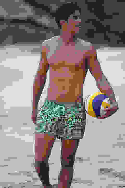 Ein Mann in Badeshorts am Strand. Unter dem Arm trägt er einen Volleyball.