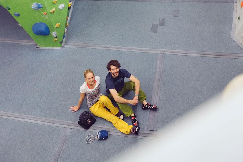 Ein Mann und eine Frau sitzen mitten in einer Boulderhalle auf dem Fussboden. Sie tragen lockere und bunte Boulderkleidung.