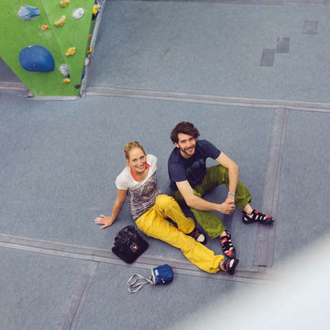 Ein Mann und eine Frau sitzen mitten in einer Boulderhalle auf dem Fussboden. Sie tragen lockere und bunte Boulderkleidung.