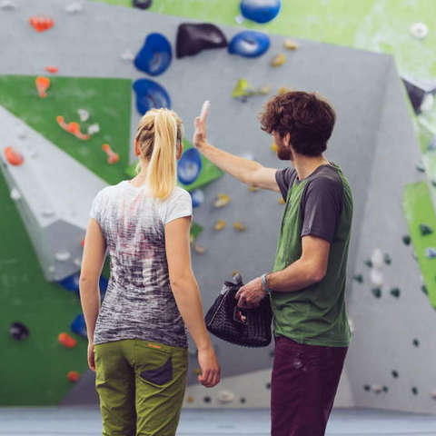 Eine Frau und ein Mann stehen vor einer Boulderwand in einer Kletterhalle und besprechen ihre Klettertaktik.