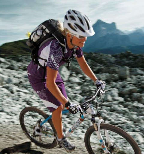 Eine Frau fährt mit einem Fahrradrucksack auf dem Rücken auf ihrem Mountainbike einen steinigen Gebirgspfad entlang.