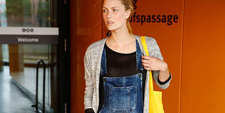 Eine Frau in Jeans Latzhose steht vor dem Eingang einer Einkaufspassage.