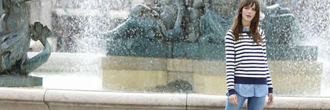 Eine dunkelhaarige Frau steht in Blue Jeans und gestreiftem Pullover vor einem Springbrunnen.