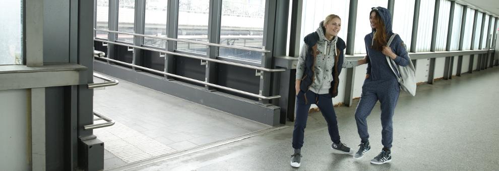 Zwei lachende Frauen in Jogginghosen stehen in einem Geb&auml;ude