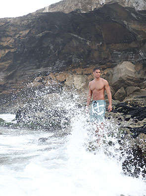 Ein Mann steht an einer Steinküste in Badehose. Das Wasser brandet gegen die Felsen.